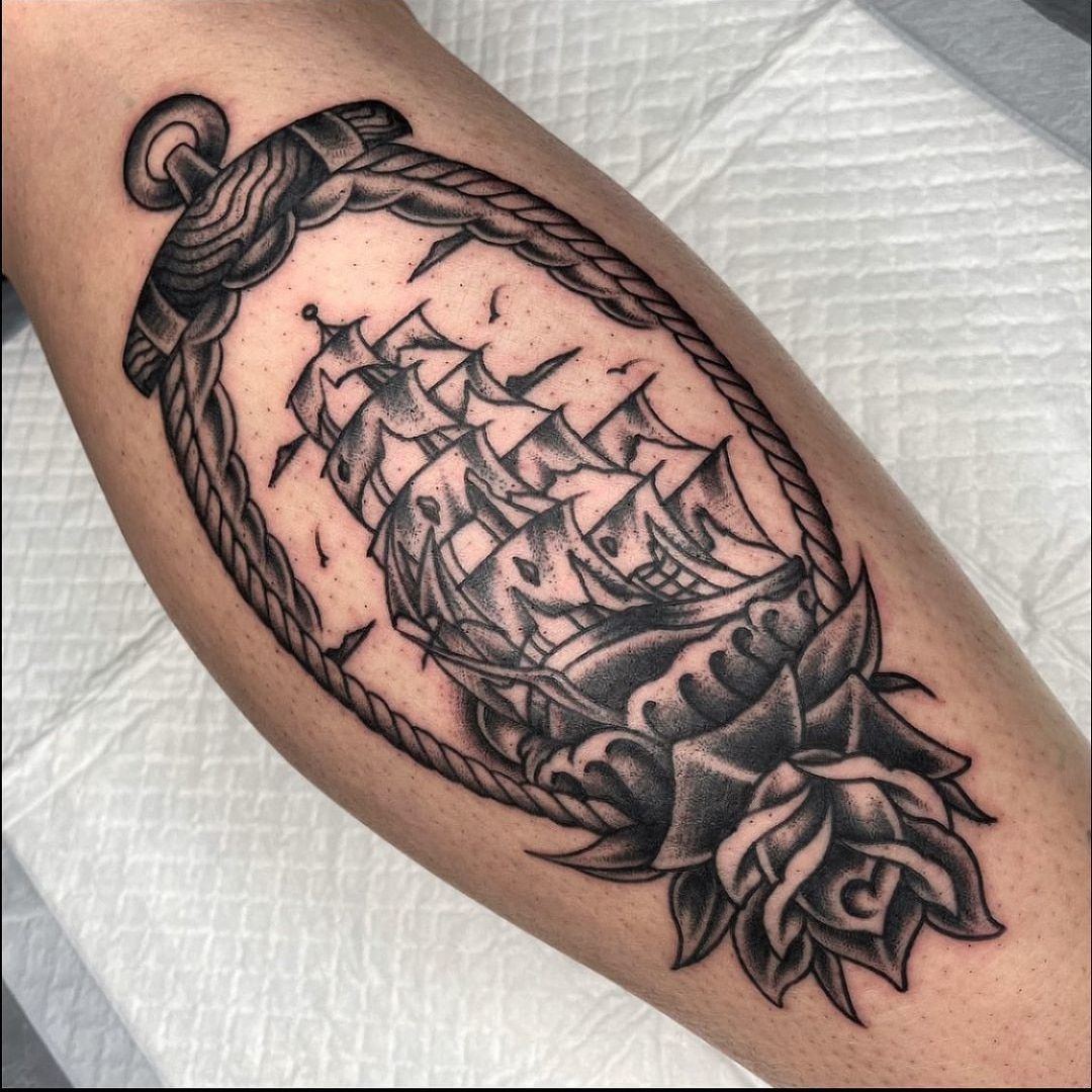 A detailed tattoo of a ship within a rope and rose design on a leg.
