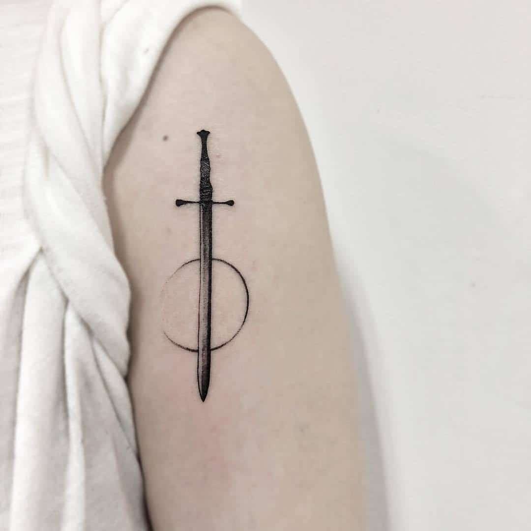 A detailed black and gray tattoo of a sword surrounded by a circle on a person's arm.